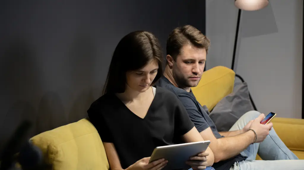 Couple sitting apart in silence, one partner looking at a phone, representing emotional disconnection and digital betrayal in relationship