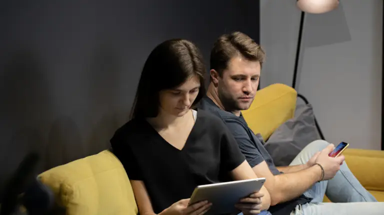 Couple sitting apart in silence, one partner looking at a phone, representing emotional disconnection and digital betrayal in relationship